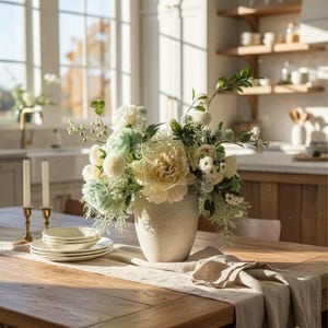 K&ouml;nnte beinhalten: Ein Holztisch ist mit einem Blumengesteck in einer strukturierten Vase gedeckt. Der Strau&szlig; zeigt cremefarbene und hellgr&uuml;ne Blumen. Zwei Kerzenst&auml;nder mit brennenden Kerzen und ein Stapel wei&szlig;er Teller stehen ebenfalls auf dem Tisch.