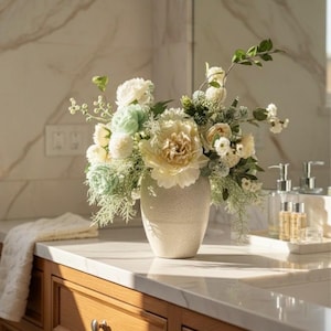 K&ouml;nnte beinhalten: Ein Blumenarrangement in einer strukturierten, cremefarbenen Vase. Der Strau&szlig; besteht aus gro&szlig;en, cremefarbenen Pfingstrosen, kleineren wei&szlig;en und hellgr&uuml;nen Bl&uuml;ten und Gr&uuml;nzeug. Die Vase steht auf einer hellen Arbeitsplatte &uuml;ber Holzschr&auml;nken.