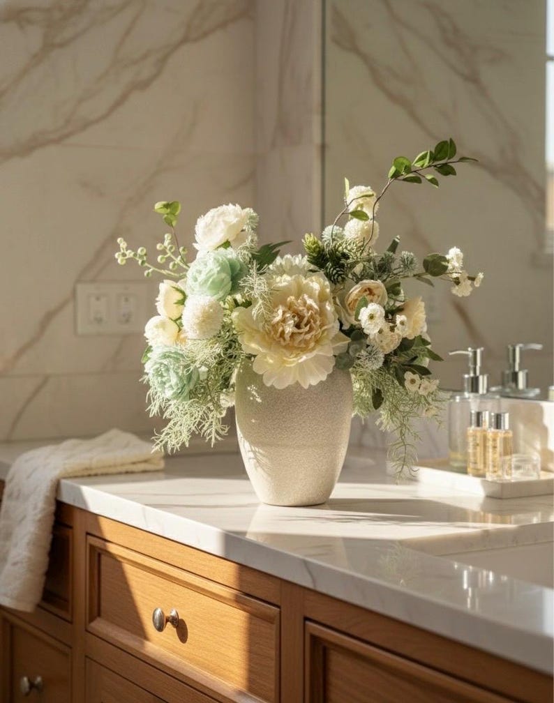 K&ouml;nnte beinhalten: Ein Blumenarrangement in einer strukturierten, cremefarbenen Vase. Der Strau&szlig; besteht aus gro&szlig;en, cremefarbenen Pfingstrosen, kleineren wei&szlig;en und hellgr&uuml;nen Bl&uuml;ten und Gr&uuml;nzeug. Die Vase steht auf einer hellen Arbeitsplatte &uuml;ber Holzschr&auml;nken.