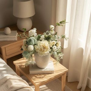K&ouml;nnte beinhalten: Eine wei&szlig;e Keramikvase mit einem Blumenarrangement aus wei&szlig;en, cremefarbenen und hellblauen Blumen steht auf einem h&ouml;lzernen Beistelltisch. Eine wei&szlig;e Lampe und B&uuml;cher befinden sich ebenfalls auf dem Tisch. Die Szene wird von nat&uuml;rlichem Licht erhellt.