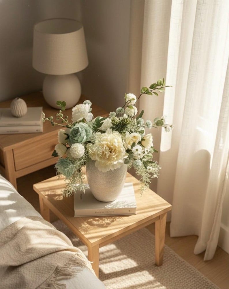 K&ouml;nnte beinhalten: Eine wei&szlig;e Keramikvase mit einem Blumenarrangement aus wei&szlig;en, cremefarbenen und hellblauen Blumen steht auf einem h&ouml;lzernen Beistelltisch. Eine wei&szlig;e Lampe und B&uuml;cher befinden sich ebenfalls auf dem Tisch. Die Szene wird von nat&uuml;rlichem Licht erhellt.