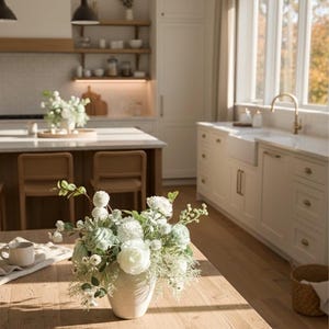 K&ouml;nnte beinhalten: Ein Holztisch zeigt eine Vase mit wei&szlig;en und hellgr&uuml;nen Blumen. Die Vase ist cremefarben und strukturiert. Eine wei&szlig;e Tasse und ein Tuch liegen ebenfalls auf dem Tisch. Die K&uuml;che hat helle Schr&auml;nke und ein gro&szlig;es Fenster.
