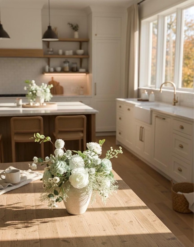 K&ouml;nnte beinhalten: Ein Holztisch zeigt eine Vase mit wei&szlig;en und hellgr&uuml;nen Blumen. Die Vase ist cremefarben und strukturiert. Eine wei&szlig;e Tasse und ein Tuch liegen ebenfalls auf dem Tisch. Die K&uuml;che hat helle Schr&auml;nke und ein gro&szlig;es Fenster.