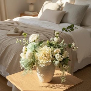 K&ouml;nnte beinhalten: Ein Blumenarrangement in einer gesprenkelten beigefarbenen Vase steht auf einem h&ouml;lzernen Beistelltisch. Es enth&auml;lt gro&szlig;e, cremefarbene gelbe Pfingstrosen, wei&szlig;e Rosen und gr&uuml;nes Laub. Im Hintergrund ein Bett mit wei&szlig;er Bettw&auml;sche und neutralfarbenen Kissen.