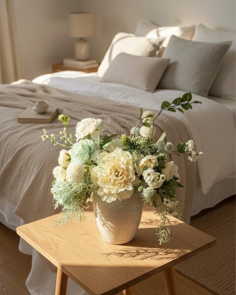 K&ouml;nnte beinhalten: Ein Blumenarrangement in einer gesprenkelten beigefarbenen Vase steht auf einem h&ouml;lzernen Beistelltisch. Es enth&auml;lt gro&szlig;e, cremefarbene gelbe Pfingstrosen, wei&szlig;e Rosen und gr&uuml;nes Laub. Im Hintergrund ein Bett mit wei&szlig;er Bettw&auml;sche und neutralfarbenen Kissen.