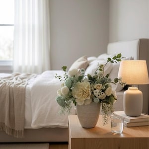 K&ouml;nnte beinhalten: Ein Schlafzimmer in neutralen T&ouml;nen mit einem Nachttisch aus Holz, auf dem eine Vase mit wei&szlig;en und gr&uuml;nen Blumen steht. Eine Lampe mit beigem Schirm steht auf dem Tisch, neben einem Glas Wasser und B&uuml;chern. Ein Bett mit wei&szlig;er Bettw&auml;sche und einer beigen Decke befindet sich im Hintergrund.
