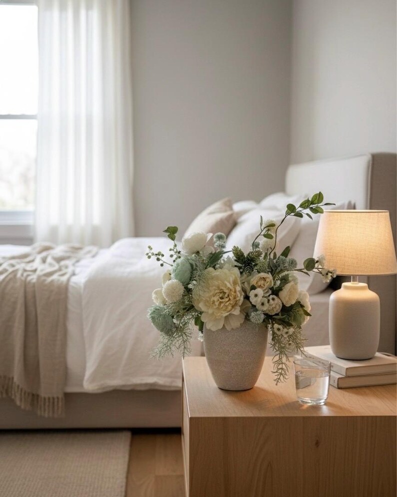 K&ouml;nnte beinhalten: Ein Schlafzimmer in neutralen T&ouml;nen mit einem Nachttisch aus Holz, auf dem eine Vase mit wei&szlig;en und gr&uuml;nen Blumen steht. Eine Lampe mit beigem Schirm steht auf dem Tisch, neben einem Glas Wasser und B&uuml;chern. Ein Bett mit wei&szlig;er Bettw&auml;sche und einer beigen Decke befindet sich im Hintergrund.