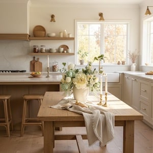 K&ouml;nnte beinhalten: Eine helle K&uuml;che mit einem Holztisch als Mittelpunkt. Der Tisch ist mit einer Vase mit wei&szlig;en und gr&uuml;nen Blumen, einem Leinenl&auml;ufer und zwei goldenen Kerzenhaltern dekoriert. Hocker und Regale aus Holz vervollst&auml;ndigen die Szene.