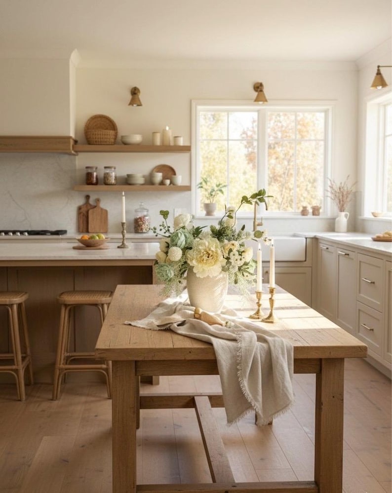 K&ouml;nnte beinhalten: Eine helle K&uuml;che mit einem Holztisch als Mittelpunkt. Der Tisch ist mit einer Vase mit wei&szlig;en und gr&uuml;nen Blumen, einem Leinenl&auml;ufer und zwei goldenen Kerzenhaltern dekoriert. Hocker und Regale aus Holz vervollst&auml;ndigen die Szene.