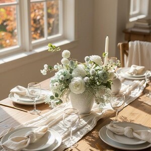 K&ouml;nnte beinhalten: Ein gedeckter Esstisch mit wei&szlig;en Tellern, Servietten und Weingl&auml;sern. Eine wei&szlig;e Vase enth&auml;lt ein Blumenarrangement mit wei&szlig;en und gr&uuml;nen Blumen. Ein wei&szlig;er Tischl&auml;ufer und goldfarbenes Besteck.