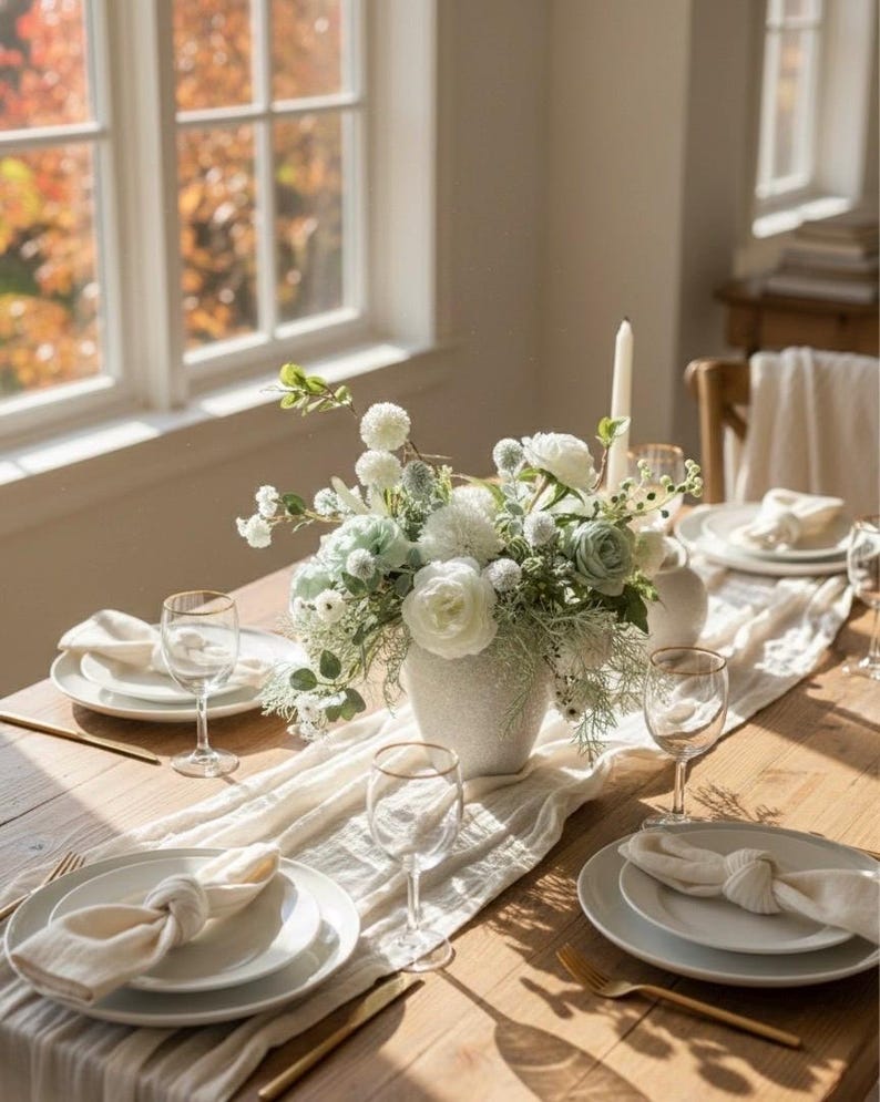 K&ouml;nnte beinhalten: Ein gedeckter Esstisch mit wei&szlig;en Tellern, Servietten und Weingl&auml;sern. Eine wei&szlig;e Vase enth&auml;lt ein Blumenarrangement mit wei&szlig;en und gr&uuml;nen Blumen. Ein wei&szlig;er Tischl&auml;ufer und goldfarbenes Besteck.