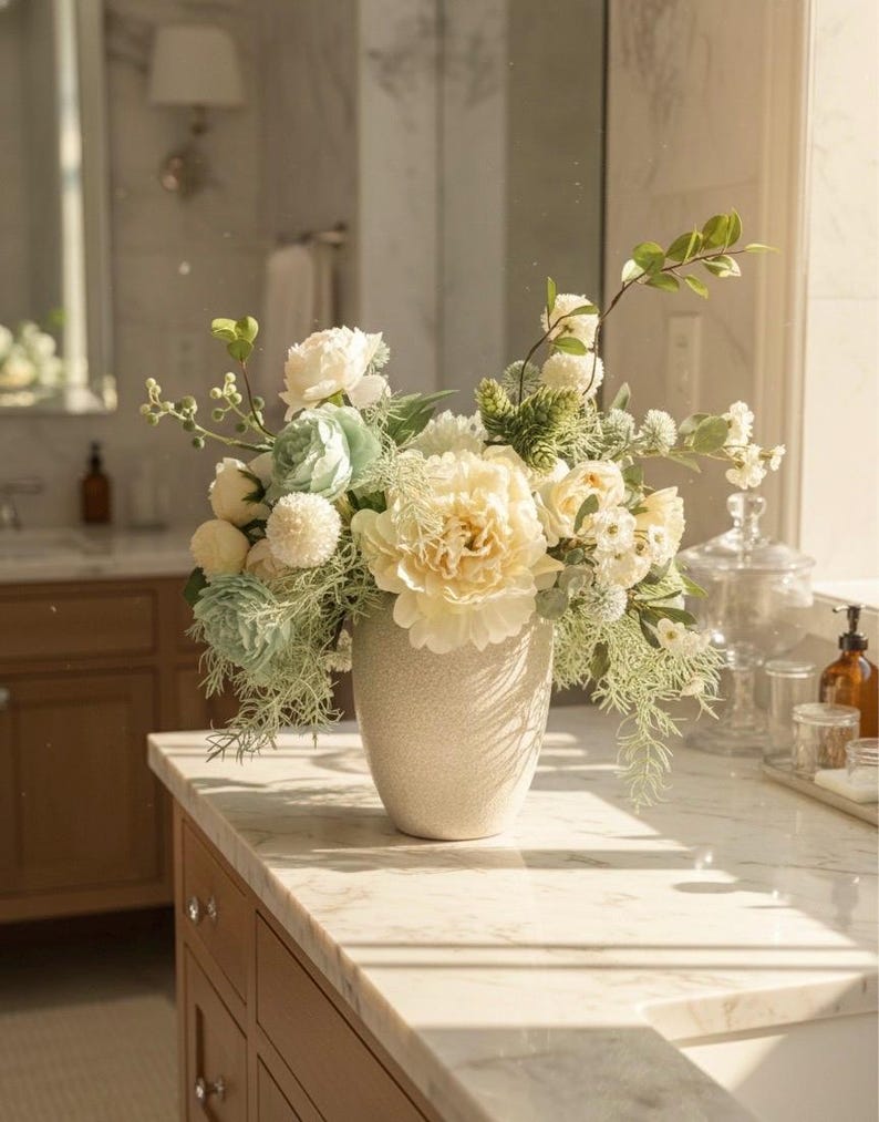 K&ouml;nnte beinhalten: Ein Blumenarrangement in einer strukturierten, cremefarbenen Vase. Der Strau&szlig; besteht aus gro&szlig;en, cremefarbenen Pfingstrosen, zarten blau-gr&uuml;nen Akzenten und verschiedenen wei&szlig;en und gr&uuml;nen Bl&auml;ttern. Die Anordnung steht auf einer Marmorarbeitsplatte.