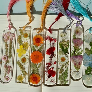 May include: Six clear resin bookmarks with colorful dried flowers and tassels. The flowers include pink, yellow, orange, and red blooms. Each bookmark has a different color tassel, including pink, gold, red, purple, and teal.