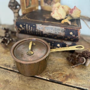 May include: A small, antique copper saucepan with a lid and brass handle. The pot sits on a wooden surface, with dried flowers and antique books in the background. The pot has a tarnished appearance.