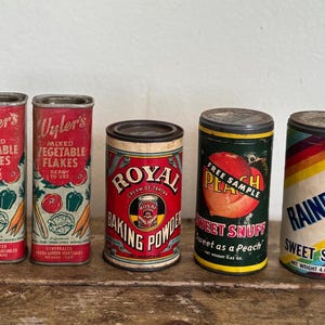 Puede incluir: Cinco latas de alimentos de metal vintage con etiquetas. Dos latas están etiquetadas como "Wyler's Mixed Vegetable Flakes", una "Royal Baking Powder", una "Sweet Snuff Sweet as a Peach" y una "Rainbow Sweet Snuff". Las latas están sobre una superficie de madera.