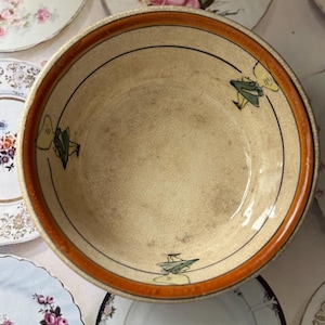 Puede incluir: Un cuenco de cerámica vintage con interior color crema y borde naranja. El cuenco presenta un diseño de líneas negras y pequeñas figuras de dibujos animados en verde y amarillo. Otros platos decorativos rodean el cuenco.