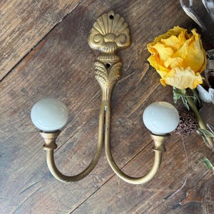 May include: A double wall hook made of brass with a decorative shell-shaped top and two curved arms. Each arm terminates in a smooth, round, white ceramic knob. The metal features a warm, metallic gold finish with visible mounting holes.