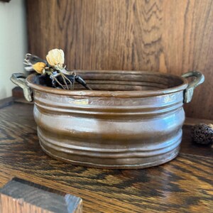 Puede incluir: Un cuenco ovalado de cobre con una textura acanalada horizontal en las paredes exteriores y dos pequeñas asas de metal en los lados opuestos. Un pequeño grupo de flores secas de color crema y amarillo descansa dentro del cuenco.