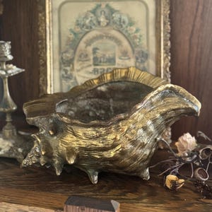 May include: A gold-colored, seashell-shaped decorative bowl with a textured surface. The bowl has a wide opening and sits on three small feet. The background includes a framed picture and wooden surfaces.