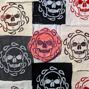 May include: A close-up of a red rubber stamp with a skull design. The stamp is surrounded by fabric squares with different coloured skull designs.