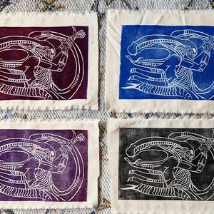 May include: Four fabric prints of a white line drawing of an alien creature on a colored background. The prints are purple, blue, black, and a lighter purple.
