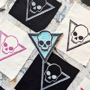 May include: Five fabric squares with a skull design printed in the center of each. The skulls are printed in different colors, including black, white, pink, and blue. The fabric squares are arranged in a grid pattern.
