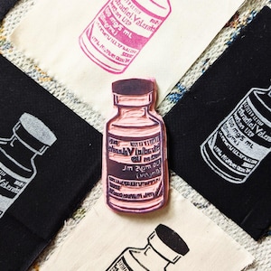 May include: A pink and white Estradiol Valerate bottle stamp is featured on black and off-white fabric squares. The stamp is in the center, surrounded by other stamped squares. The text on the bottle reads "Estradiol Valerate" and "100 mg/5 mL".