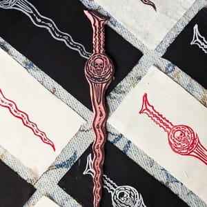 May include: A pink wooden carving of a sword with a skull design. The sword is surrounded by white fabric and black fabric with the same design.