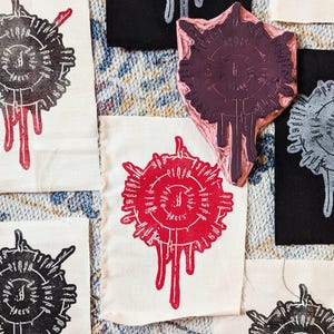 Peut inclure: Gros plan sur des carrés de tissu avec un motif circulaire répétitif à l'encre rouge, noire et grise. Le motif comprend du texte et des éléments dégoulinants. Un tampon avec le même motif est également visible.