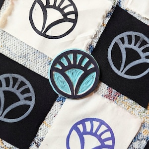 May include: Four different fabric squares with a stylized sun design printed on them. The designs are in black, gray, blue, and teal. The teal design is a wooden stamp.