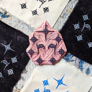 May include: A pink rubber stamp with a detailed design of a face with eyes and a starburst pattern. The stamp is surrounded by fabric squares with printed designs of eyes and stars.