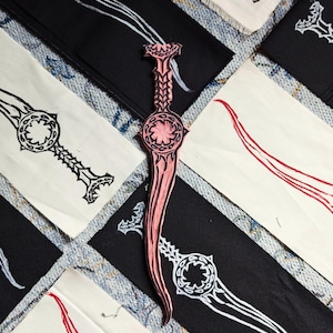 Może przedstawiać: Drewniana stempla w kształcie różowego miecza z misternymi detalami, otoczona białą tkaniną z czarno-białymi wzorami mieczy.