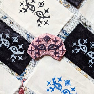 May include: A close-up of a pink rubber stamp with a design of two eyes surrounded by stars. The stamp is on a white fabric surface with other fabric squares with the same design in black and blue.