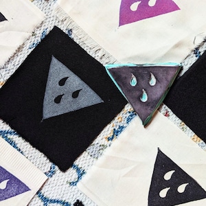 Pode incluir: Um close-up de três quadrados de tecido com um design de triângulo impresso em cada um. Os triângulos são de diferentes cores, incluindo roxo, preto e cinza. Os triângulos têm três formas de lágrima dentro. Os quadrados de tecido estão sobre uma superfície com um padrão branco e amarelo.