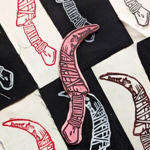 May include: A pink rubber stamp of a curved knife with a black outline and a detailed design of wrapped bandages. The stamp is on a black and white fabric background.