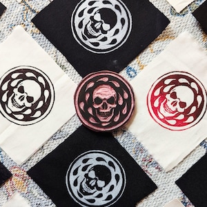 May include: An assortment of fabric squares and a circular stamp, all displaying a skull motif. The skull is encircled by a decorative pattern. The central stamp is pink, with the squares alternating between black and white fabric.
