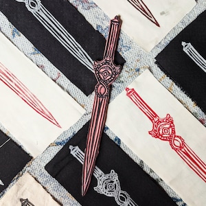 May include: A red wooden sword with intricate details and a black and white striped pattern. The sword is lying on a white fabric surface with other swords in the background.