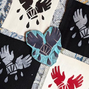 May include: Four fabric squares featuring a design of two hands clasped together, dripping with red, black, silver, and blue paint. The design is a block print, and the fabric squares are arranged in a circle.