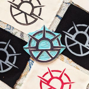 May include: A close-up of a wooden printing block with a blue and black design of a sun with rays. The block is surrounded by fabric squares with the same design printed in black, grey, and red.