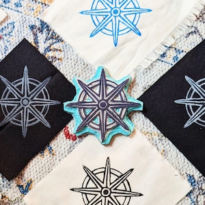 May include: An assortment of fabric squares and a stamp featuring a compass rose design. The squares are black and off-white, with the compass rose printed in blue and gray. The stamp itself is a light blue color.