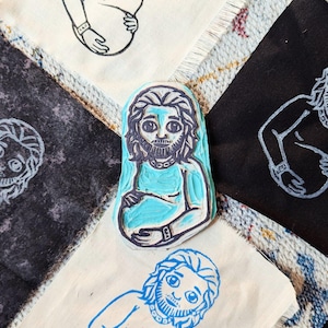May include: A blue rubber stamp featuring a stylized illustration of a person with a beard, holding their belly. The stamp is surrounded by fabric squares with similar printed designs in black and blue ink. The overall theme is related to art and crafting.
