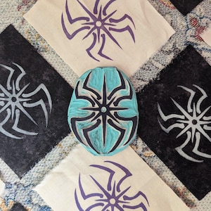 May include: A teal blue wooden block with a carved starburst design. The block is surrounded by four fabric squares with the same design printed in black, white, and purple.