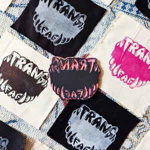 Pode incluir: Quadrados de tecido com as palavras "TRANS" e "FAG" impressas em uma fonte estilizada. Os quadrados são pretos, brancos e rosa. Um carimbo preto com as palavras "SMART" e "DAZE" está no centro.