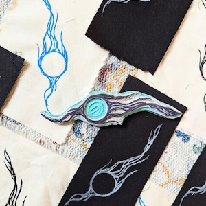 May include: An overhead shot of a wooden stamp with a teal and brown design, placed on a patchwork of fabric. The stamp has a circular center with a flame-like design. Surrounding the stamp are black fabric pieces with white flame designs and blue flame designs on cream fabric.