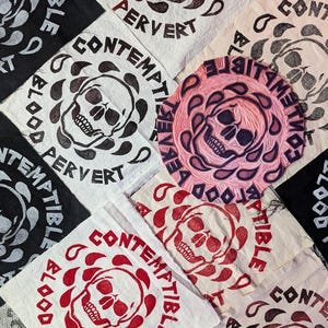Puede incluir: Primer plano de varios parches de tela con un diseño de calavera y huesos cruzados. Los parches están impresos con las palabras "Contemptible" y "Pervert" en tinta negra y roja. Los parches están dispuestos en un patrón aleatorio sobre un fondo blanco.