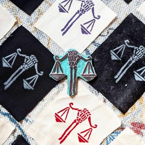 May include: A collection of fabric patches featuring a skeleton hand holding a scale design. The patches are in various colours, including blue, red, and grey. The patches are on a textured fabric background.