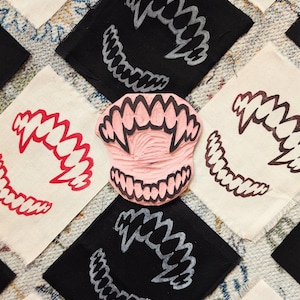 May include: A pink rubber stamp of a set of teeth is surrounded by black and white fabric squares with teeth designs. The teeth designs are in red, white, and silver. The fabric squares are arranged on a patterned surface.