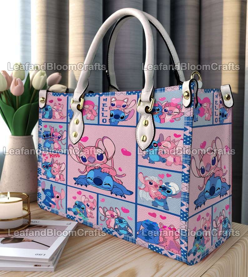 Op de afbeelding: Een rechthoekige handtas met een herhalend patroon van blauwe en roze stripfiguren in verschillende houdingen met hartjes. De tas heeft witte handvatten, goudkleurige hardware en een rasterontwerp met de tekst HELLO en Kiss.