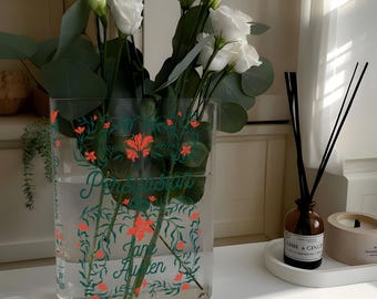 May include: A clear glass vase with a floral design featuring green leaves and orange flowers. The vase is filled with water and white flowers. The vase has the text "Persuasion" and "Jane Austen" printed on it.