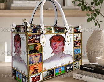 May include: A structured handbag featuring a collage of David Bowie imagery, including the Aladdin Sane album cover. The bag has white top handles, gold-tone hardware, and a white base color with various rectangular panels displaying portraits.